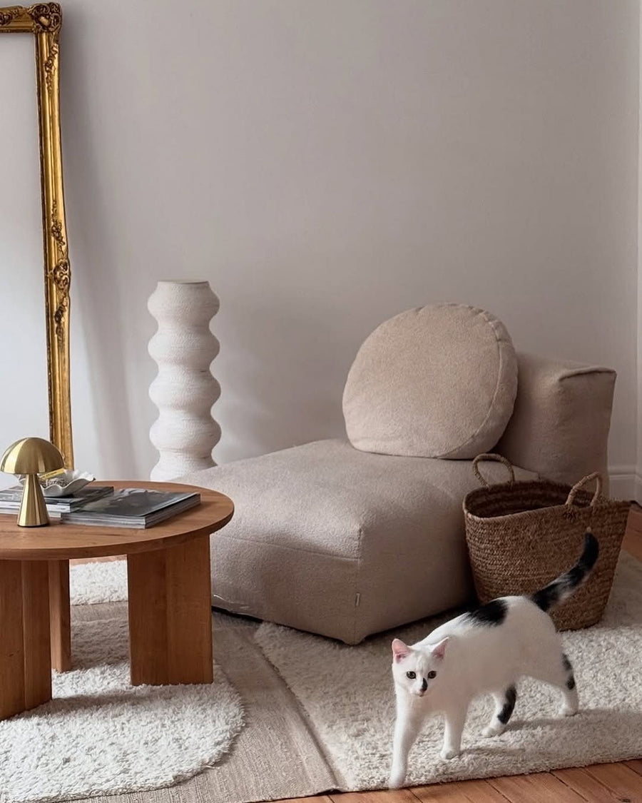 Chauffeuse Cloud en tissu beige avec coussin rond, table basse en bois, miroir doré et un chat dans un salon minimaliste