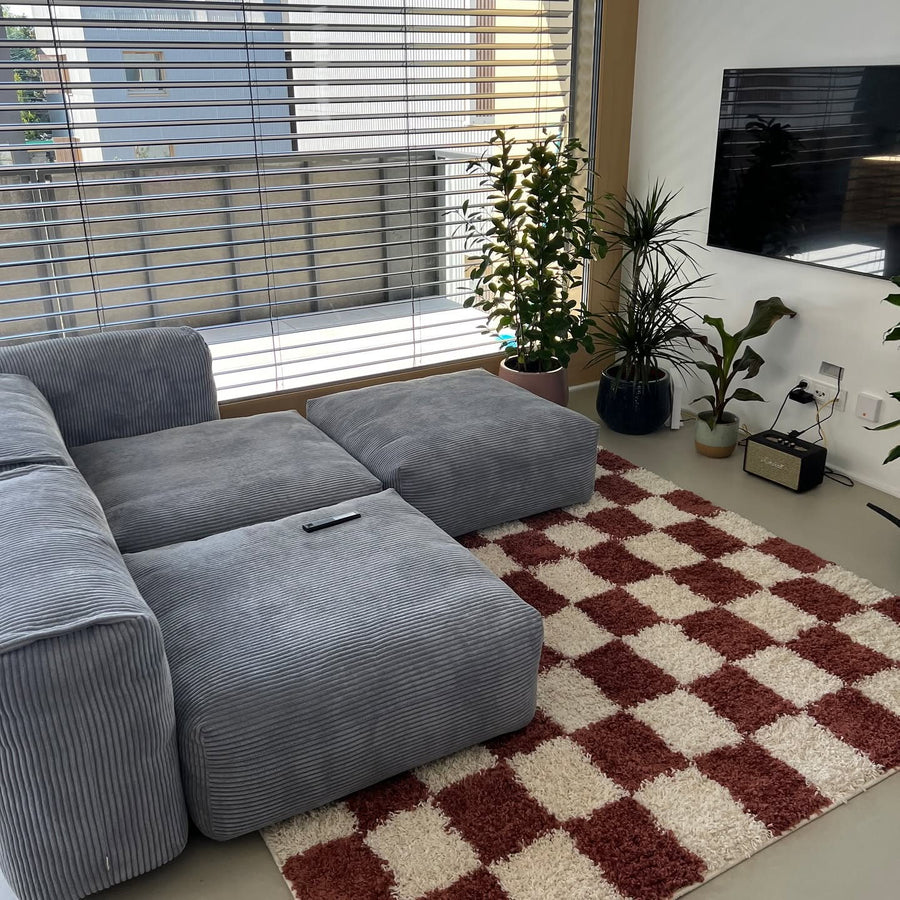 Canapé modulable Cloud en velours côtelé gris-bleu devant une baie vitrée, tapis à damier bordeaux et plantes vertes