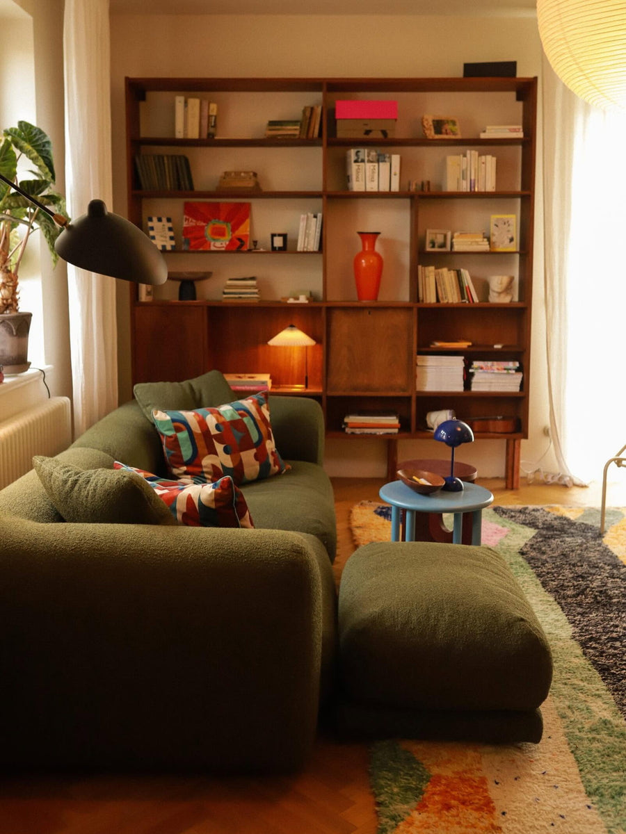 Canapé modulable Lazy en tissu vert olive avec pouf devant une grande bibliothèque en bois, lampe et table d’appoint bleue