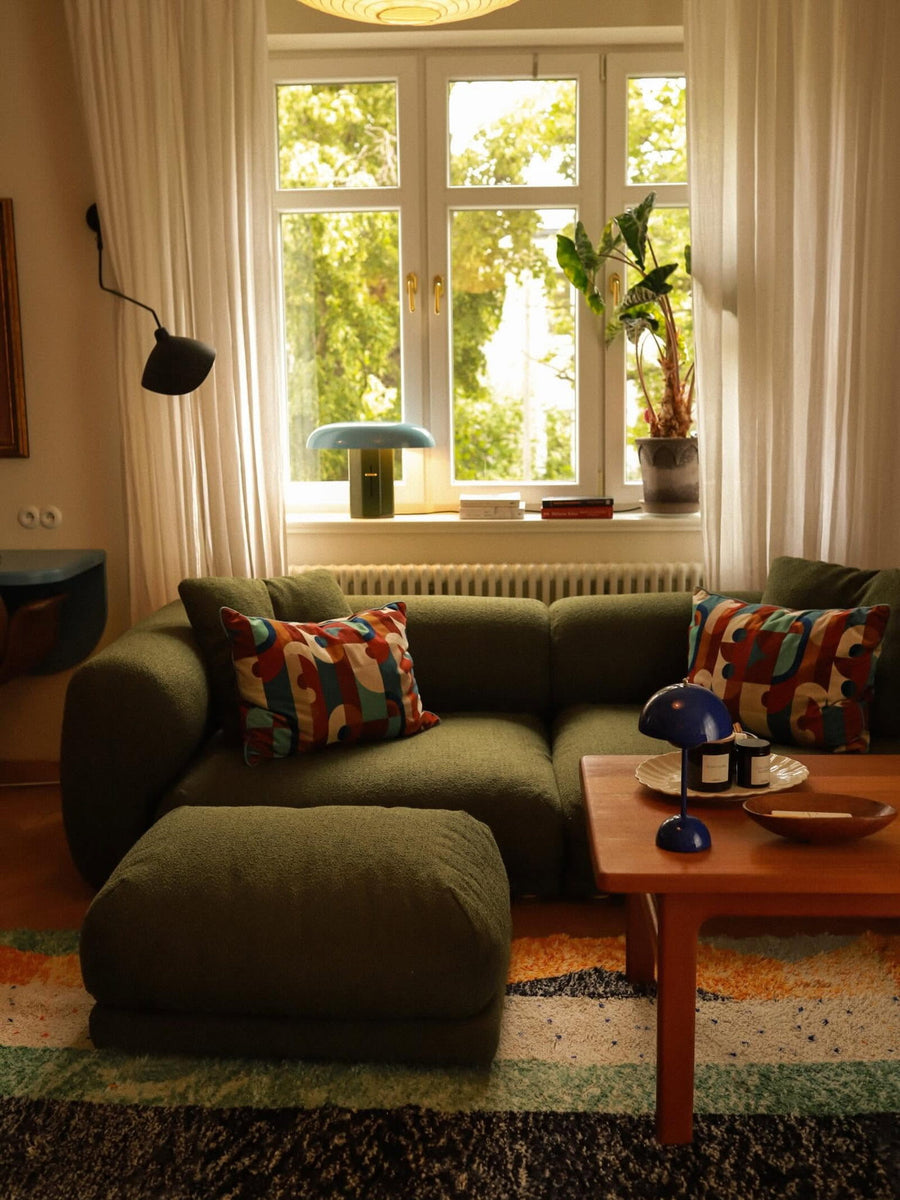 Canapé modulable Lazy en tissu vert olive avec pouf et coussins à motifs devant une fenêtre, table basse en bois