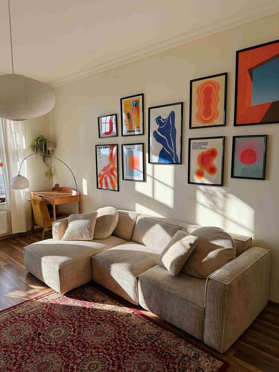 Canapé modulable Slay en velours côtelé beige sous un mur de cadres colorés, lampe arc et tapis persan